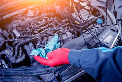 A mobile auto repair person wearing red gloves holds a blue cloth, checking a dipstick in a sunlit car engine bay, conveying a sense of maintenance and care.