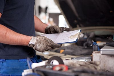 A Vegas Auto Repair mechanic in gloves examines a checklist clipboard near an open car hood in a driveway, surrounded by tools